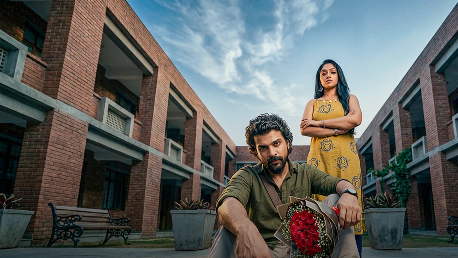 Man seated in a courtyard holding a bouquet of red roses, with a woman in a yellow dress standing behind him and arms crossed.